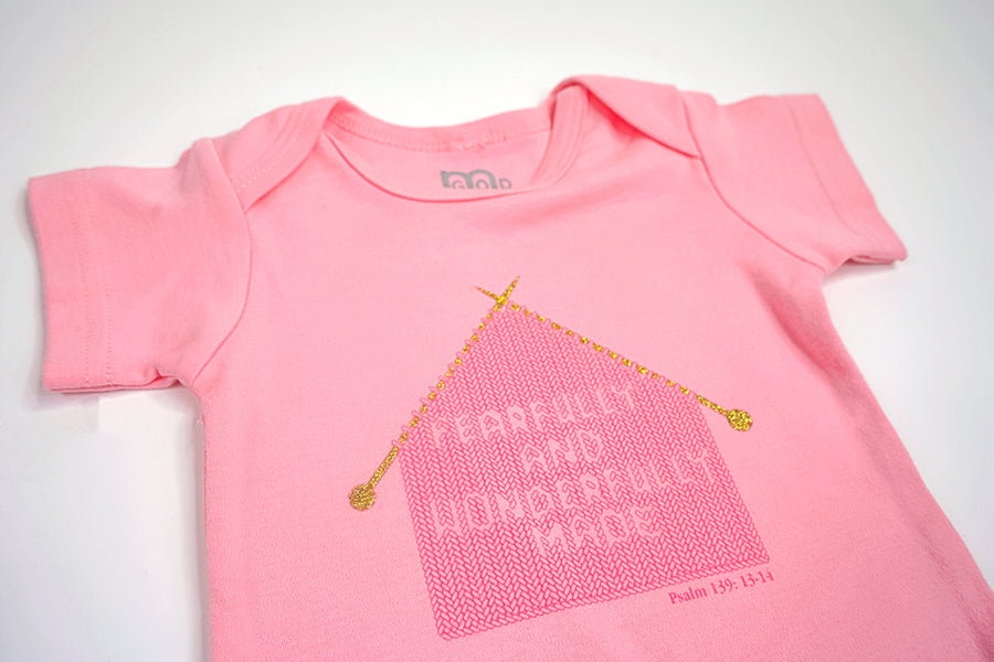 Pink Infant Onesie with "Fearfully and Wonderfully Made" printed on front and woven tag GODinme logo at left leg.
